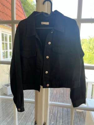 Svart jeansjacka från Gina Tricot - Snygg svart jeansjacka från Gina Tricot med klassisk krage, silverfärgade knappar och två bröstfickor med lock. Jackan har en rak och något kortare passform, perfekt att slänga över en hoodie eller t-shirt.