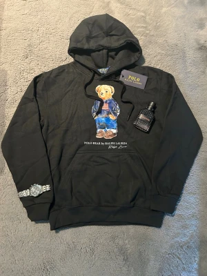 Svart hoodie Polo Bear  - Svart hoodie med Polo Bear-trycket framtill. Hoodien har huva med dragsko och en klassisk känguruficka. Materialet är mjukt och bekvämt, perfekt för en avslappnad stil. Snygg detalj med björnen i jeans och stickad tröja.