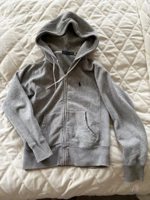 Grå zip-hoodie från Polo Ralph Lauren - Klassisk grå hoodie med dragkedja från Polo Ralph Lauren. Har huva med vita snören, två fickor fram och broderad logga på bröstet. Mjuk bomullsblandning och ribbade muddar vid ärmslut och nederkant. Perfekt för en chill och stilren look. Har använts av en 13/14 årig kille som är 168cm lång