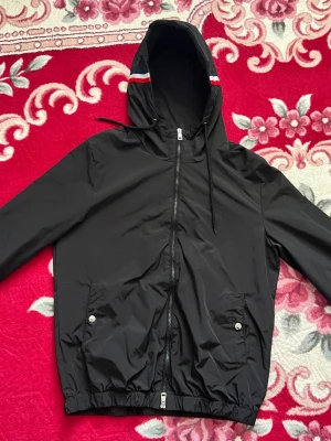 Svart Moncler vindjacka med huva - Svart vindjacka från Moncler med dragkedja framtill, två fickor med tryckknappar och elastisk nederkant. Jackan har huva med röd, vit och blå rand samt Moncler-logga på ärmen. Perfekt för blåsiga dagar och snygg streetstil. Äkta hoodie