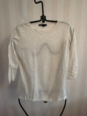 Vit prickig topp från Mango - Vit topp från Mango med små svarta prickar över hela tyget. Modellen har rund halsringning och trekvartsärm. Tillverkad i mjukt bomullsmaterial som känns skönt mot huden. Perfekt för en avslappnad och stilren look.