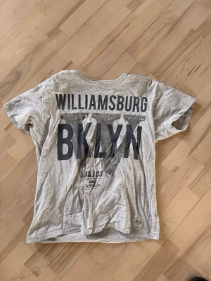 Grå T-shirt med Williamsburg-tryck - Grå T-shirt från H&M med stort svart tryck på ryggen där det står 'WILLIAMSBURG BKLYN'. T-shirten har rund halsringning och är tillverkad i mjuk bomull. Perfekt för en avslappnad streetstyle-look.