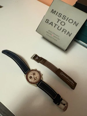 Omega x Swatch MoonSwatch Saturn - Omega x Swatch Speedmaster MoonSwatch Mission to Saturn med beige boett, brun bezel och detaljer, samt mörkblått läderband med vita sömmar. Extra brunt textilband medföljer. Ikonisk kronografdesign och tachymeter på ringen. C O P I A!! Efter marnknads band och sitt band följer med nytt batteri med