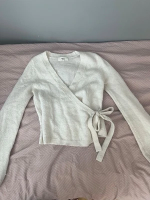 Vit omlottstickad tröja från Gina Tricot - Säljer en vit stickad omlotttröja från Gina Tricot med långa ärmar och knytband i sidan. Tröjan har ribbstickade detaljer vid ärmslut och nederkant, och är gjord i ett mjukt material som känns skönt mot huden. Perfekt för att skapa en snygg lager-på-lager look.