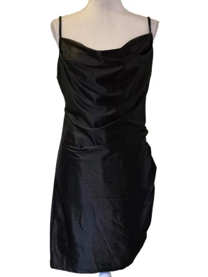 Svart klänning i storlek L Nypris 399 kr - Smala axelband med snyggt fall fram. Glatt glansigt satin tyg. Rynkning en sida nedtill. Svart foder. Material 100 % polyester foder, 95 % polyester och 5 % elastan ytterdel. Bystbredd ca 47 cm, midjebredd ca 42 cm och längd ca 80 cm från ovandel. 1179/68