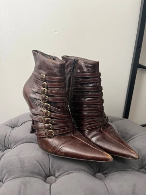 Bruna stövletter med spännen - Unika bruna stövletter i skinn med spetsig tå och höga klackar. Snygga remmar med flera metallspännen längs sidan ger en cool och edgy look. Dragkedja på insidan för enkel påtagning. Perfekta för dig som vill sticka ut med din stil.
