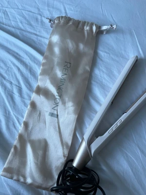 Plattång Remington - Säljer en stilren plattång från Remington i vitt och guld med digitala knappar. Kommer med en snygg, glansig förvaringspåse i ljusbeige tyg. Perfekt för att styla håret snabbt och smidigt. Har inte använt max 5 gng
