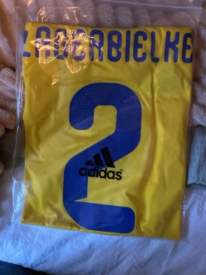 Gul Adidas fotbollströja nr 2 - Säljer en gul fotbollströja från Adidas med blått tryck, nummer 2 och namnet 'Lagerbielke' på ryggen. Tröjan har sportig design och är tillverkad i ett lätt och ventilerande material, perfekt för träning eller match.