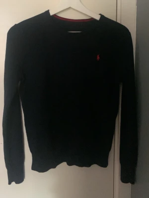 Svart stickad tröja från Ralph Lauren xs/s 165/170 cm - Pris kan diskuteras. Klassisk mörkblå stickad tröja från Ralph Lauren med rund halsringning och röd broderad logga på bröstet. Tröjan har långa ärmar och ribbade muddar vid ärmslut och nederkant. Perfekt för en stilren och enkel look. Den har ett pyttelitet hål på armen som knapps syns och går att sy