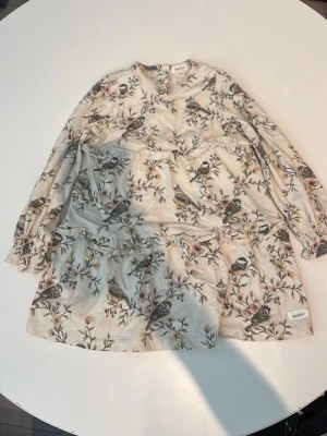 Blommig klänning från Newbie - Supersöt ljus klänning från Newbie med blommigt och fågelmönster. Klänningen har lång ärm, rund hals och volangdetalj nedtill. Perfekt för vår och sommar, tillverkad i mjuk bomull för extra komfort.