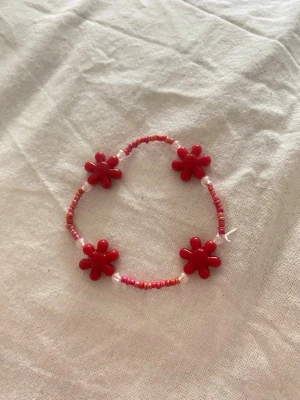 Rött pärlarmband med blommor - Sött armband med små röda och rosa pärlor samt fyra större röda blommor i plast. Perfekt accessoar för att ge en färgklick till din outfit. Armbandet är elastiskt och passar de flesta handleder. Egengjord