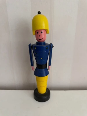 Träfigur soldat blå och gul Brio - Säljer en charmig träfigur föreställande en soldat med blå uniform, gula detaljer och hjälm samt svart bälte. Figurerad med ett leende ansikte och enkel design. Perfekt som dekoration eller samlarobjekt.