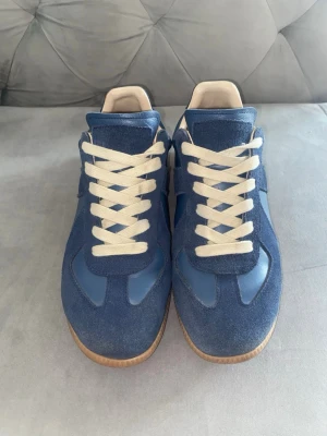 Maison Margiela - Snygga blå sneakers med ovandel i mocka och läder, vita skosnören och klassisk gummisula. Skorna har en retroinspirerad design med låg profil och beige detaljer vid häl och plös. Endast skorna ingår och ingen original skokartong. Perfekta för dig som gillar stilrena och tidlösa sneakers.