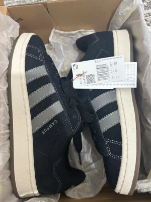 Svarta Adidas Campus sneakers - Snygga svarta Adidas Campus sneakers med klassiska tre vita ränder på sidan och vit sula. Ovandelen är i mocka och skorna har svarta skosnören. Perfekt för dig som gillar retrostil och streetwear. Diskret Adidas-logga och Campus-tryck på sidan.