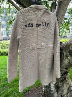 Beige stickad kappa Odd Molly - Snygg beige stickad kappa från Odd Molly med stora knappar framtill, två fickor och dekorativ detalj i ryggen. Kappan har en klassisk krage och är perfekt för lager-på-lager-stil. Broderad logga på ryggen ger en unik touch.