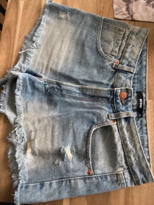 Blå jeansshorts med fransar från Bik Bok - Snygga blå jeansshorts från Bik Bok med slitna detaljer och fransig kant. Klassisk femficksmodell med knapp och dragkedja. Perfekta för varma dagar och enkel att matcha med olika toppar.