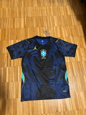 Blå Brasilien fotbollströja  - Snygg blå och svart fotbollströja , med gula detaljer. Tröjan har korta ärmar, rund hals och är tillverkad i ett lätt, ventilerande material perfekt för träning eller match.