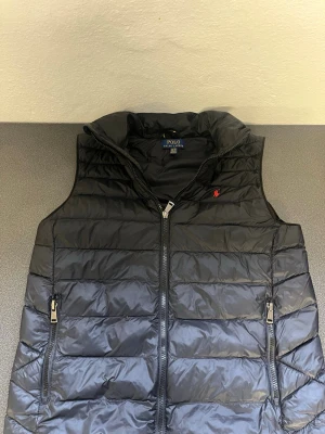 Svart dunväst från Polo Ralph Lauren - Snygg svart dunväst från Polo Ralph Lauren med quiltad design och dragkedja framtill. Västen har två fickor med dragkedja och en liten röd logga på bröstet. Perfekt för lager-på-lager och streetstyle.