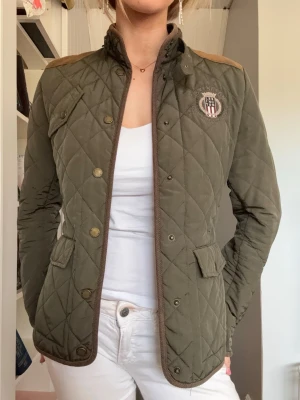 Quiltad grön jacka från Lexington - Snygg quiltad jacka i mörkgrönt från Lexington med bruna detaljer och emblem på bröstet. Jackan har knappar framtill, två fickor med lock och axelpartier i kontrastfärg. Perfekt för vår, höst & sommar!!💕