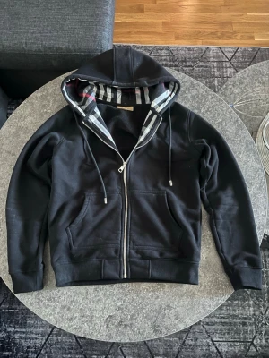 Burberry Hoodie Svart - Svart hoodie med dragkedja från Burberry. Klassisk modell med huva och dragsko, samt fickor framtill. Insidan av huvan har Burberrys ikoniska rutmönster i svart, vitt och rött. Perfekt för en stilren och avslappnad look.
