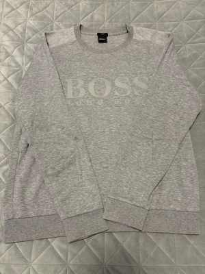 Grå sweatshirt från Hugo Boss - Snygg grå sweatshirt från Hugo Boss med stor logga framtill. Tröjan har rund halsringning, ribbade muddar vid ärmslut och nederkant. Perfekt för en avslappnad och stilren look. Materialet är mjukt och bekvämt.