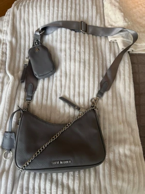 Grå axelväska från Steve Madden - Snygg grå axelväska från Steve Madden i stilren design. Väskan har en bred axelrem, silverfärgad kedjedetalj och ett litet avtagbart fack. Tillverkad i konstläder med struktur och logga framtill. Perfekt för dig som vill ha en trendig och praktisk väska.