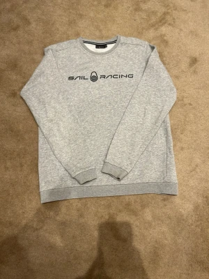 Grå sweatshirt från Sail Racing - Snygg grå sweatshirt från Sail Racing med svart logotyptryck på bröstet. Tröjan har rund halsringning, långa ärmar och ribbade muddar vid ärmslut och nederkant. Tillverkad i mjukt bomullsmaterial för en bekväm känsla. Pris kan diskuteras!! Vid minsta fundering är det bara att höra av sig!