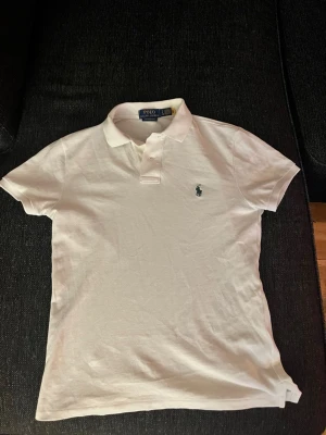Vit pikétröja från Polo Ralph Lauren - Klassisk vit pikétröja från Polo Ralph Lauren med broderad logga på bröstet. Tröjan har krage, korta ärmar och knäppning framtill. Modellen är custom slim fit och tillverkad i bomull för en skön känsla.