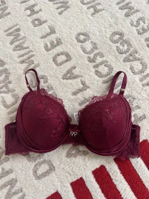 Vinröd spets-bh med rosett - Snygg vinröd bh med vadderade kupor och vacker spetsdetalj över hela framsidan. Dekorerad med en liten rosett och strassdetalj mellan kuporna. Justerbara axelband och hakknäppning bak. Perfekt för dig som gillar feminina detaljer.