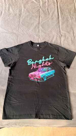 Svart t-shirt Clique Bangkok Nights - Svart t-shirt från Clique med tryck i neonfärger som föreställer en bil och texten 'Bangkok Nights' på bröstet. Aldrig använd 