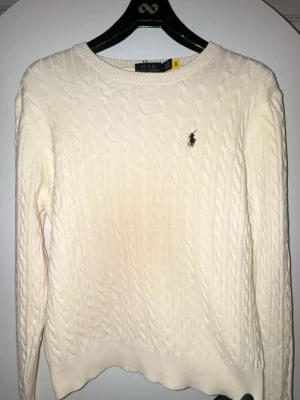 Vit kabelstickad tröja Polo Ralph Lauren - Kabelstickad vit tröja från Polo Ralph Lauren med rund halsringning och den klassiska broderade loggan på bröstet. Tröjan har långa ärmar och ribbade muddar vid ärmslut och nederkant. Perfekt för lager på lager och stilrena outfits. Passar M