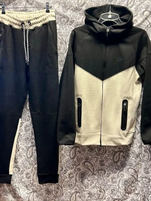 Nike svart och vit tracksuit set - Tvådelat tracksuit set från Nike i svart och vit. Setet består av en hoodie med dragkedja, huva och två fickor med dragkedja samt matchande byxor med dragsko i midjan och sidofickor. Tillverkad i mjukt sweatshirtmaterial, perfekt för chill dagar.