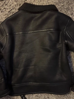 Svart Rebel skinnjacka med fuskpäls - Snygg från Rebel, svart skinnjacka med bred krage och fuskpälsfoder. Jackan har dragkedja framtill, två snedställda fickor med dragkedja och detaljerade sömmar. Perfekt för dig som vill ha en cool och varm look i vinter.