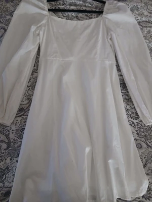 Vit långärmad klänning med puffärm - Stilren vit klänning med långa puffärmar och fyrkantig ringning. Klänningen har en lätt glansig yta och är sydd i ett slätt material som ger en elegant känsla. Passar perfekt för dig som gillar en klassisk och trendig look.