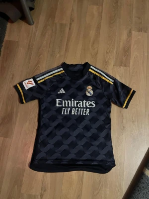 Real Madrid matchtröja Adidas svart - Snygg svart Real Madrid matchtröja från Adidas med gulddetaljer och klubbmärke på bröstet. Tröjan har korta ärmar, rund hals och trycket 'Emirates Fly Better' på framsidan. Perfekt för fotbollsträning eller att bära till vardags.
