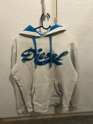 Vit hoodie från Diesel med blå detaljer - Säljer en vit hoodie från Diesel med blå broderad logga på bröstet, blå snören och blått innerfoder i huvan. Klassisk känguruficka framtill och mjukt material som känns skönt mot huden. Perfekt för dig som gillar streetwear och vill sticka ut lite extra.