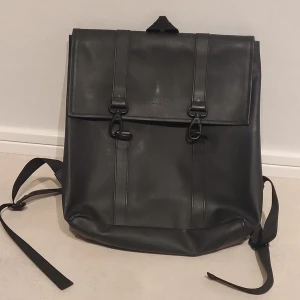 Svart ryggsäck i konstläder - Stilren svart ryggsäck från Rainsi slätt konstläder. Ryggsäcken har två justerbara axelband, lock med två spännen framtill och en enkel, fyrkantig form. Perfekt för dig som gillar minimalistisk design och vill ha plats för det viktigaste.