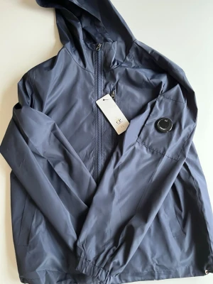 Blå vindjacka från C.P. Company - Snygg blå vindjacka från C.P. Company med huva och dragkedja framtill. Jackan har en ikonisk linsdetalj på ärmen och elastiska muddar. Tillverkad i lätt, vattenavvisande material som passar perfekt för blåsiga dagar.