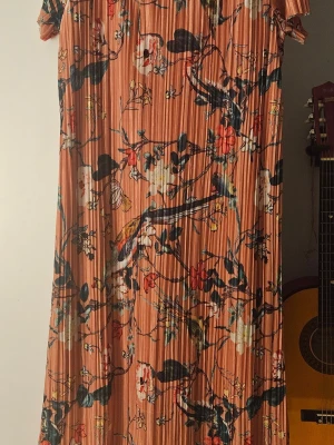 Blommig plisserad klänning i orange - Säljer en plisserad klänning i en varm orange ton med blommigt mönster i vitt, rött, blått och grönt. Klänningen har korta ärmar och en rak, lös passform som ger en avslappnad vibe. Perfekt för dig som gillar färg och mönster!