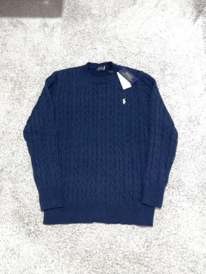 Mörkblå kabelstickad tröja Polo Ralph Lauren - Mörkblå kabelstickad tröja från Polo Ralph Lauren med rund halsringning och klassisk vit broderad logga på bröstet. Tröjan har ribbade muddar vid ärmslut och nederkant och är tillverkad i mjukt material. Perfekt för lager på lager under kalla dagar.