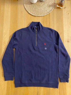 Mörkblå half-zip tröja Polo Ralph Lauren - Mörkblå långärmad tröja från Polo Ralph Lauren med half-zip och ståkrage. Tröjan har det klassiska röda Polo-logot på bröstet och är tillverkad i mjukt bomullsmaterial. Perfekt för en avslappnad men stilren look.