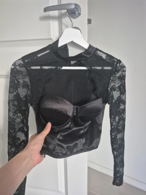 Svart spetsig croppad topp - Snygg svart croppad topp med satinkorsett och transparenta spetsärmar. Toppen har lång ärm och hög hals med en liten rosett i nacken. Perfekt för dig som vill sticka ut med en edgy och trendig look. Tar bort annons söndag!