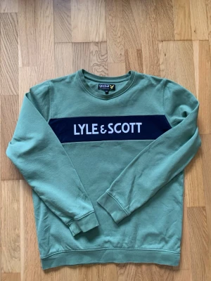 Grön sweatshirt från Lyle & Scott - Snygg grön sweatshirt från Lyle & Scott med rund hals och ett mörkblått band över bröstet där loggan är tryckt i vitt. Tröjan har ribbade muddar vid ärmslut och nederkant. Perfekt för en avslappnad och trendig stil. Pris kan diskuteras. Tveka inte på att höra av er vid eventuella frågor!