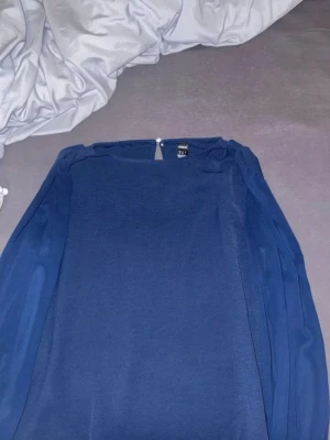 Mörkblå blus  - Snygg mörkblå blus med raka linjer och långärmade transparenta ärmar. Blusen har en enkel knappdetalj i nacken. Flow material 