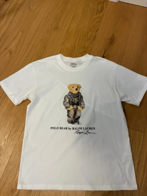 Vit Polo Bear t-shirt från Ralph Lauren - Vit t-shirt från Ralph Lauren med ikoniskt Polo Bear-motiv på bröstet. Björnen har på sig en svart skinnjacka och tröja med tigertryck. Klassisk passform och rund hals. Perfekt för dig som gillar streetstyle och vill ha något unikt i garderoben.