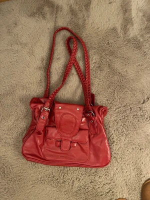 Röd handväska med flätade axelband - Snygg röd handväska i skinnimitation med flätade axelband och silverfärgade detaljer. Väskan har ett stort huvudfack, en ficka framtill med lock och spänne samt dekorativa sömmar. Perfekt för dig som vill ha en färgklick till outfiten.