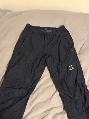 Haglös byxor - Säljer ett par svarta skalbyxor från Haglöfs med dragkedjor vid fickorna och logga på låret. Byxorna har resår i midjan och är tillverkade i ett lätt, vindtätt material. Perfekta för utomhusaktiviteter och äventyr.