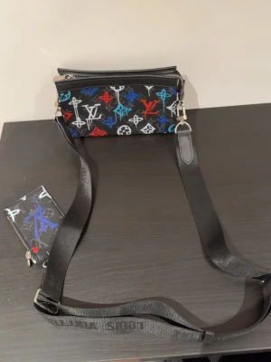 Louis Vuitton axelväska med färgglatt mönster - Snygg axelväska från Louis Vuitton i svart med färgglada LV-loggor i blått, rött och vitt. Väskan har justerbar axelrem med Louis Vuitton-tryck och medföljande liten plånbok. Perfekt för dig som vill sticka ut med en trendig accessoar.