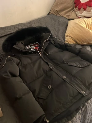 Svart pufferjacka från Moose Knuckles - Säljer en svart pufferjacka från Moose Knuckles med stor huva och svart fuskpäls. Jackan har flera fickor med lock och dragkedja, samt detaljer i metall. Perfekt för kalla vinterdagar och riktigt snygg streetstil.