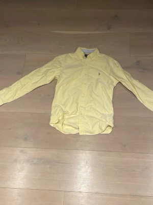 Gul skjorta från Ralph Lauren - Klassisk gul skjorta från Ralph Lauren med lång ärm och knappstängning framtill. Skjortan har en broderad logga på bröstet och är tillverkad i bomull. Perfekt för en stilren och avslappnad look.